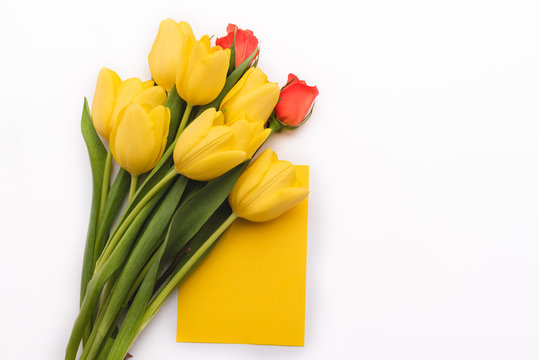 Yellow tulips and roses arranged on the left of the frame. In the middle there is a card with the possibility to save the text. Flowers lie on white isolated background. Frame with free, copy space.
