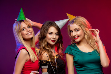Portrait of three amazing, cute girls in fashion dresses and party caps, holding sparkler or Bengal light celebrating the New year, Christmas or Birthday party, standing over colorful background
