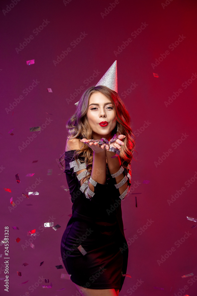 Wall mural Event, party, Birthday, Christmas or New Year celebrating concept. Young pretty brunette woman in black dress and birthday hat is laughing.. Colorful studio portrait with bright background. Copy space