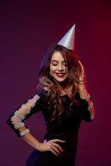 Event, party, Birthday, Christmas or New Year celebrating concept. Young pretty brunette woman in black dress and birthday hat is laughing.. Colorful studio portrait with bright background. Copy space