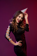 Event, party, Birthday, Christmas or New Year celebrating concept. Young pretty brunette woman in black dress and birthday hat is laughing.. Colorful studio portrait with bright background. Copy space