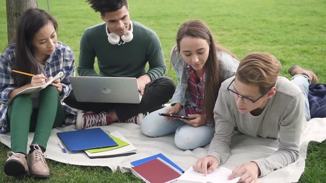Cooperation team is sitting and studying near university campus while computer networking and writing all important information in notebooks. Smart cheerful friends are learning and sufring to improve