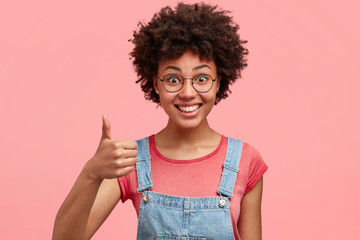 Isolated shot of attractive young woman with curly hair, smiles positively, keeps thumb raised, expresses approval, like, agreement, wears casual t shirt and denim dungarees, isolated over pink wall