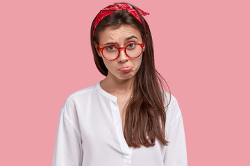 Lonely dissatisfied brunette lady purses lower lip, looks unhappy, discontent with bad choice of friend, wears red headband, white shirt, models against pink background. Negative emotions concept