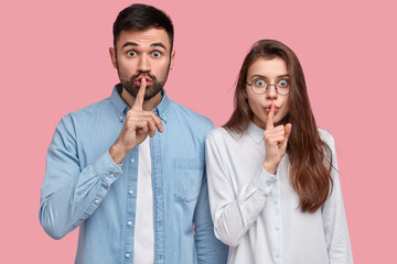 Secret and silence concept. Shot of surprised Caucasian lady and guy hold fore fingers over lips,...