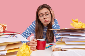 Unhappy woman with dissatisfied expression purses lower lip, wears striped clothes, drinks coffee or tea, has to fill tax form, works with documentation, isolated over pink background. Paper work