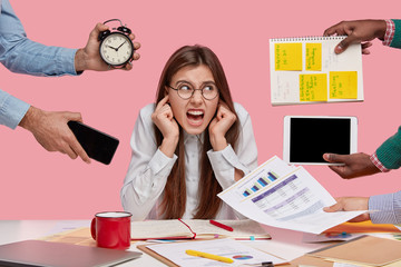 Photo of irritated woman with annoyed expression, plugs ears, ignores colleagues who have different...
