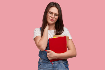 Horizontal shot of dissatisfied young woman keeps hand on neck, has painful feelings, carries red notepad, tired after long hours of working, isolated over pink background. Fatigue student or pupil