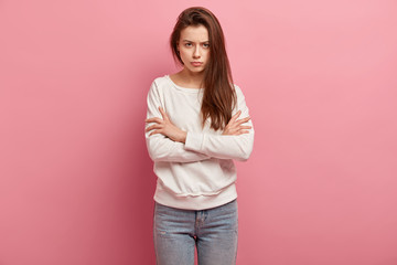 Irritated young exasperate woman keeps arms crossed, strongly dissatisfied with situation, has frowned eyebrows, demonstrates anger, wears casual jumper and jeans, stands against pink background