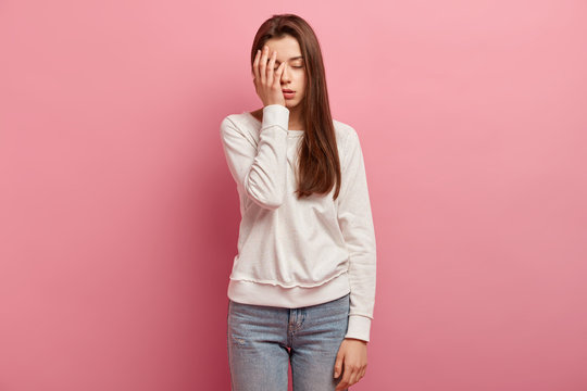 Young woman has tired expression, covers part of face with palm, closes eyes, dressed in casual clothes, wants to sleep, isolated over pink background, feels bored and displeased, has problem