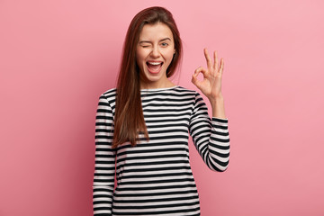 Photo of cheerful young woman blinks eye, makes okay gesture, demonstrates her agreement, feels...