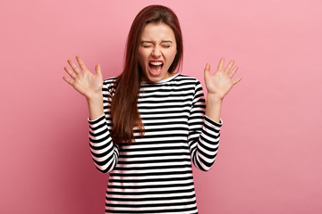 Overemotive pleased Caucasian woman exclaims happily, shows palms, wears casual striped jumper, isolated over pink background, closes eyes, reacts on sudden rumor. People and feelings concept