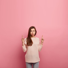 Sorrowful dissatisfied woman purses lower lip, indicates with both index fingers, shows free space above, dressed in casual sweater, has dark hair, isolated over pink background with blank place