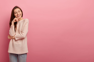 Good looking young girl keeps hand under chin, focused aside with cheerful dreamy expression, feels pleased, dressed in casual sweater, models against pink studio wall, empty space for your promotion