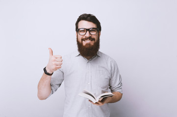 Portrait of smart cheerful  bearded man in shirt, holding a book and shows thumb up