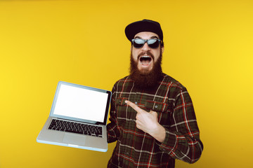 Handsome excited bearded young guy in sunglasses and hat, holding laptop and pointing at white...