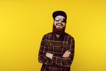 Obraz premium Portrait of smiling bearded guy wearing black hat and sunglasses, crossing hands over yellow background