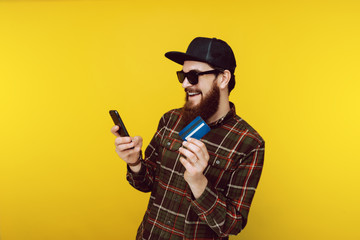 Cool hipster in sunglasss and checkered shirt is using mobile whilw showing his new crdit card