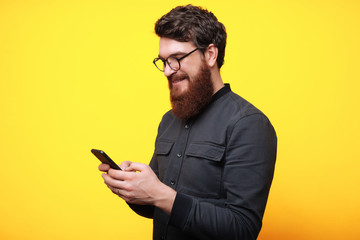 Portrait of cheerful smart beardedguy  using his smartphone over yellow background