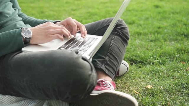 Close up hands of african college student surfing internet sitting on grass. Young man using laptop computer for browsing, communication and studying.