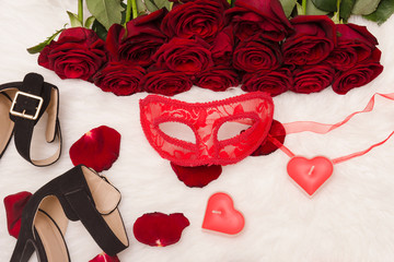 Red carnival mask, a bouquet of red roses, black shoes with heels, candles in the shape of a heart and scattered petals on the white fur.