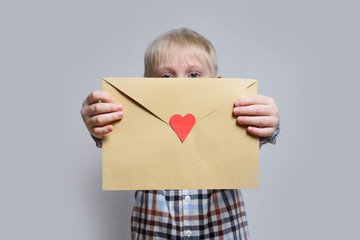 Little blond boy holds an envelope with heart on light background.