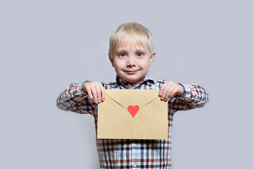 Smiling little blond boy holds an envelope with heart on light background.