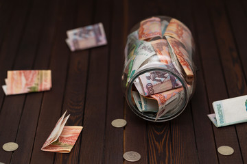 Glass jar full of money tipped over on its side spilling money on wooden background. 