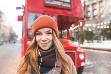 Close-up portrait of a positive girl in headphones looks at the camera on the back of a red bus.Cute smiling girl in headphones against the backdrop of urban winter landscape.Winter walk