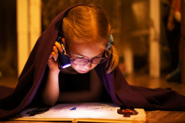 Little blonde girl 7 years old with glasses reading the book under the blanket with little flashlight at dark night time