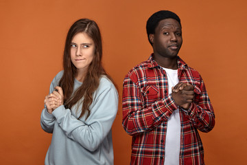 Horizontal image of interracial couple pulling tricks on each other, posing isolated against blank orange studio wall background, looking suspicious, planning trick or prank, rubbing hands
