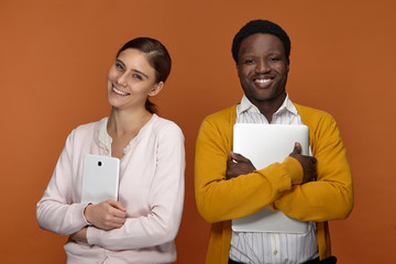 Stylish young mixed race team of two coworkers using electronic gadgets for work, positive cheerful white female carrying digital tablet and handsome smiling black male embracing laptop computer