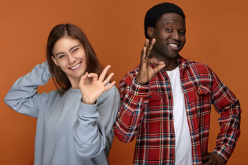 Isolated shot of overjoyed positive cute young European woman with cute broad smile posing in studio with her attractive cheerful black boyfriend both winking at you and showing okay gestures