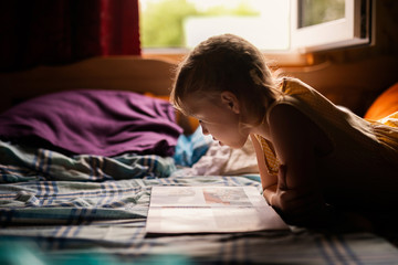 Fototapeta na wymiar Cute little blonde girl with braids reading the fairy tales book and laying on the bed near the window