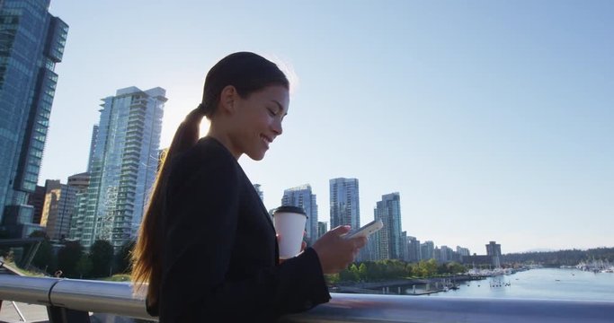 Woman in Vancouver harbour downtown looking at phone - businesswoman in city - businesswoman taking coffee break enjoying coffee. Multicultural model. Young urban professional. SLow motion.