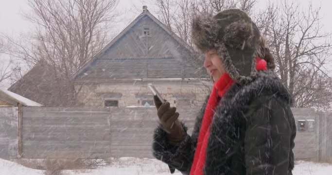 Young happy woman in red scarf and black artificial fur coat with smartphone in her hands walks through heavy snow along rural road. Bad weather, storm warning, village, winter, snow