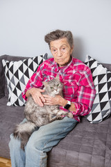 Obraz premium very old senior Caucasian grandmother with gray hair and deep wrinkles is sitting at home on the couch in jeans and a red checkered shirt with a gray fluffy shaggy cat in her arms. Pensioner and pet