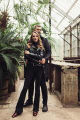 Young gorgeous woman in total black clothes dreamily looking in camera while handsome man embracing her behind. Beautiful couple spending time together in amazing greenhouse