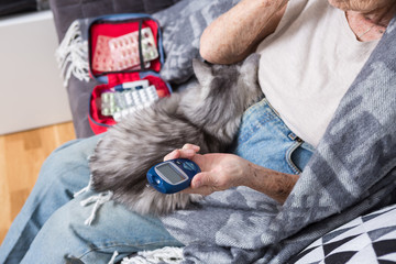 Theme old man and diabetes. Close-up of a hand. Senior Caucasian woman at home on the couch measures level of glucose in the blood with a medical device using glucometer. In the hands of apet cat