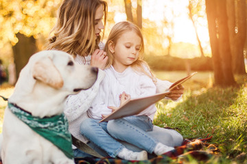 Happy beautiful mother and daughter with dog labrador is reads book sitting in the park.