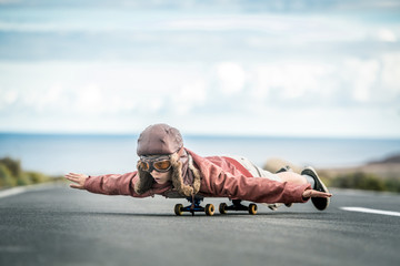 Beautiful child lying skateboard flies vintage pilot suit with hat leather jacket  mask takes off from road with sea horizon smiles amused landing taxiing departure arrival. Concept image