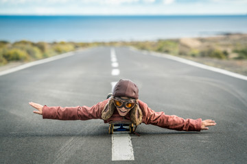 Beautiful child lying skateboard flies vintage pilot suit with hat leather jacket and mask takes off from road central line with sea horizon smiles amused landing taxiing departure arrival. Concept - Powered by Adobe