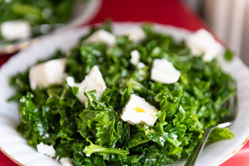 Macro closeup of kale white feta cheese green vegan vegetarian salad in restaurant with olive oil and plate on red table