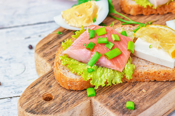 Sandwich with herring and tuna fillets with fresh green salad, onion ring, a quarter of eggs, home cheese, piece of lemon, micro greens and scallions on the wooden desk