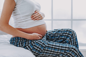 Pregnant lady in pajamas touching her tummy in light bedroom