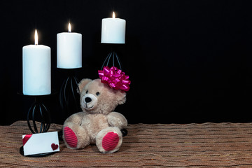 teddy bear with pink bow on head, white candles perched on black candle holders on mesh place mat and wooden table with card and dark background. 