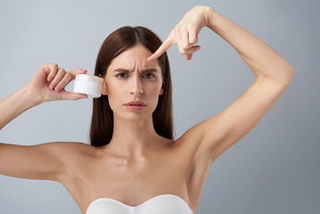 Annoyed young woman with cosmetic product showing skin problems on her forehead