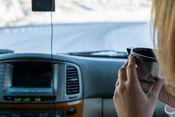 The girl in the passenger seat of the car drinking hot tea from a mug of a thermos, looking on the mountain winter road outside the car window