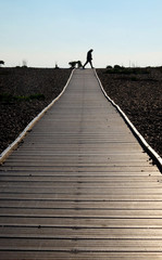 Silhouetted sixty year old man walking across the end of a long wooden pathway with a diminishing perspective