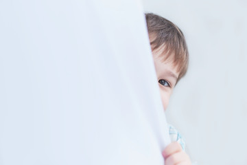 funny boy three years old looks out from behind the curtains, plays hide and seek